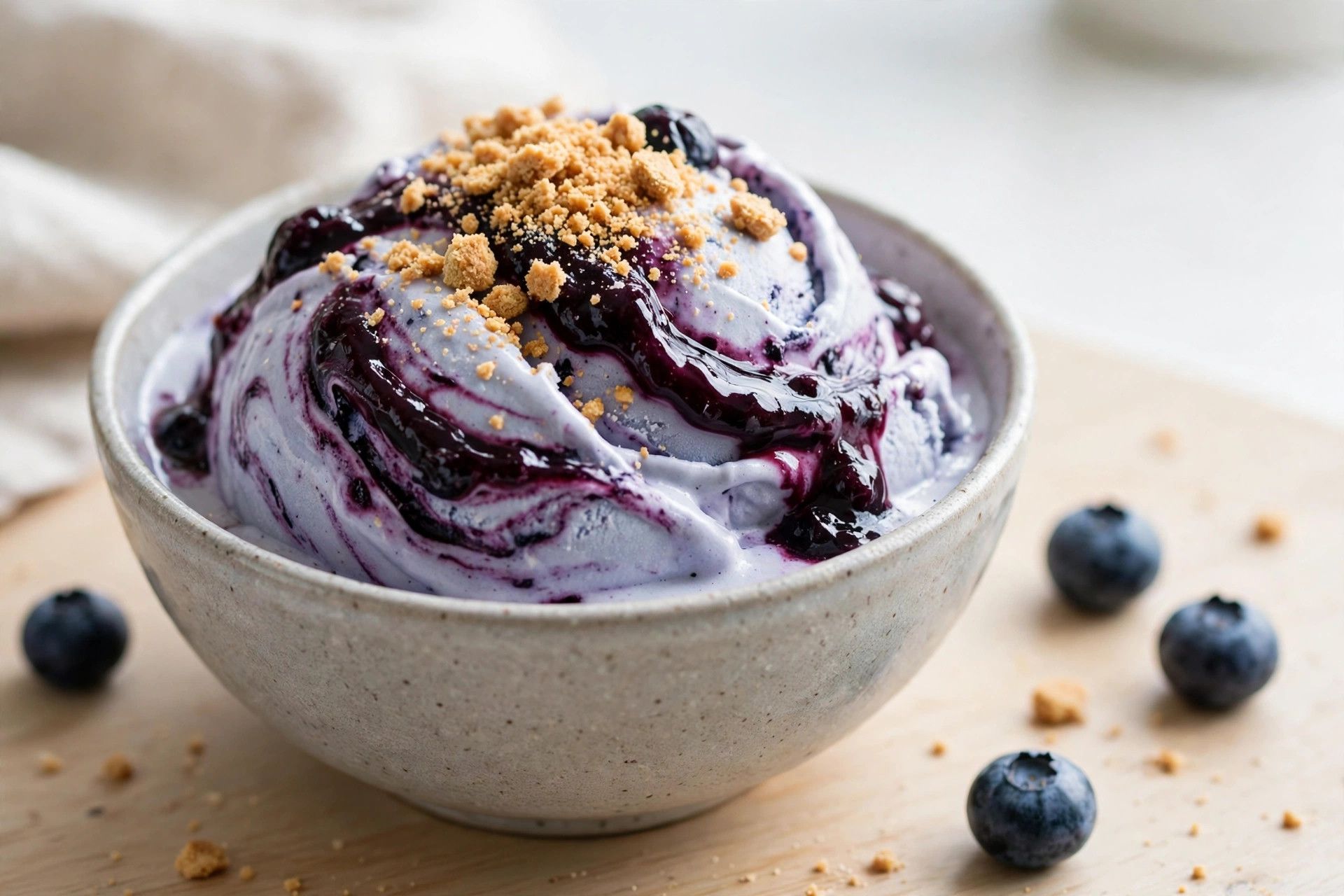 A bowl of blueberry cobbler protein ice cream with deep purple blueberry compote swirls and graham cracker crumble, on a light kitchen table