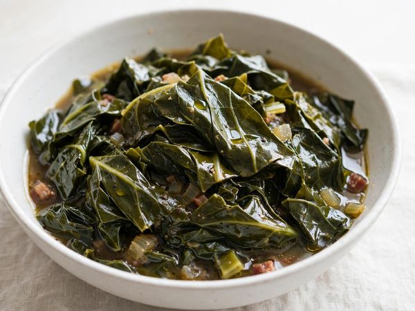 A bowl of tender southern collard greens, silky dark green leaves glistening in savory broth, on a light table
