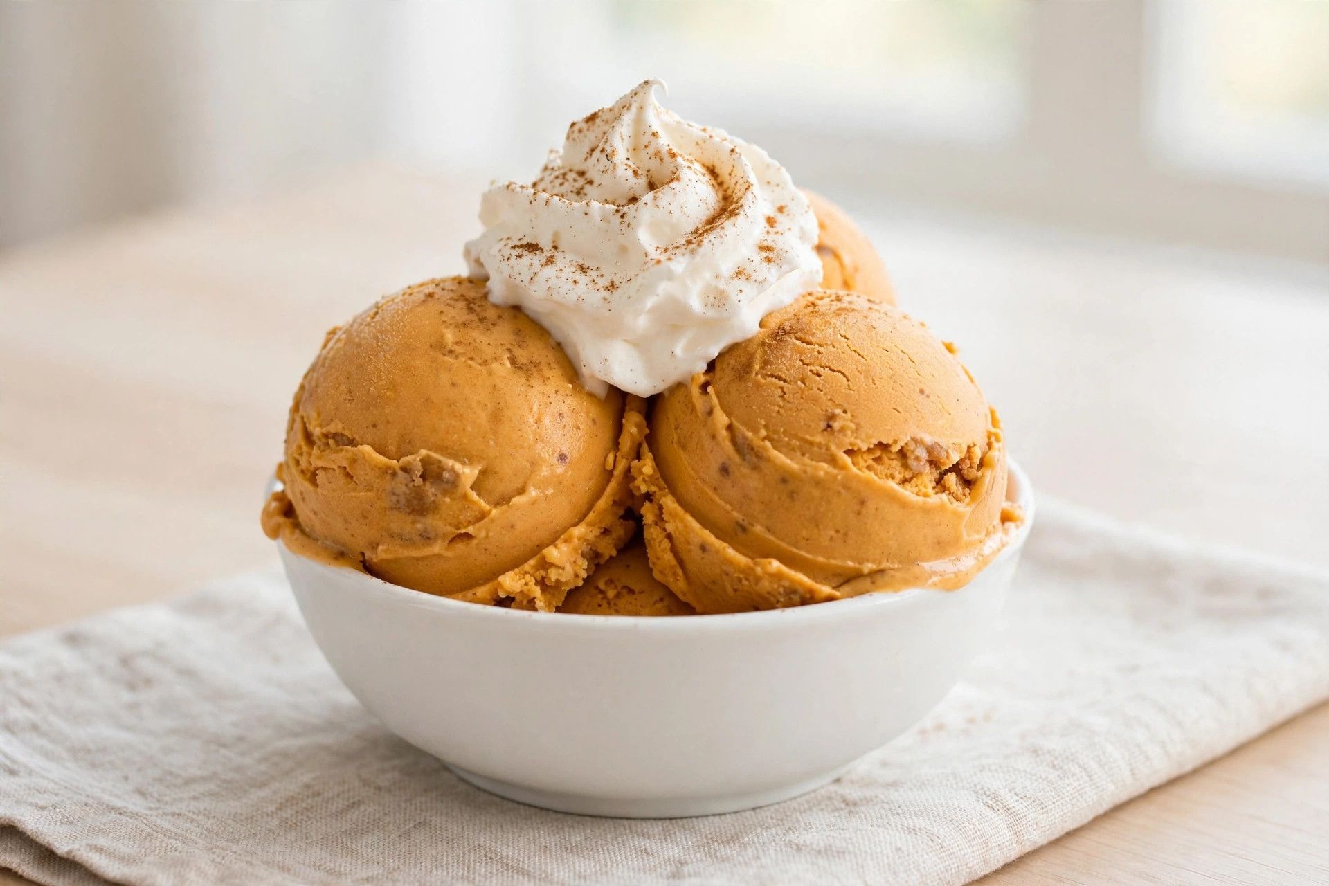 A bowl of pumpkin pie ninja creami scoops, deep orange and creamy, topped with whipped cream and cinnamon on a light table