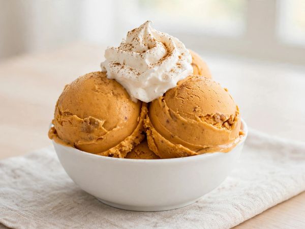 A bowl of pumpkin pie ninja creami scoops, deep orange and creamy, topped with whipped cream and cinnamon on a light table