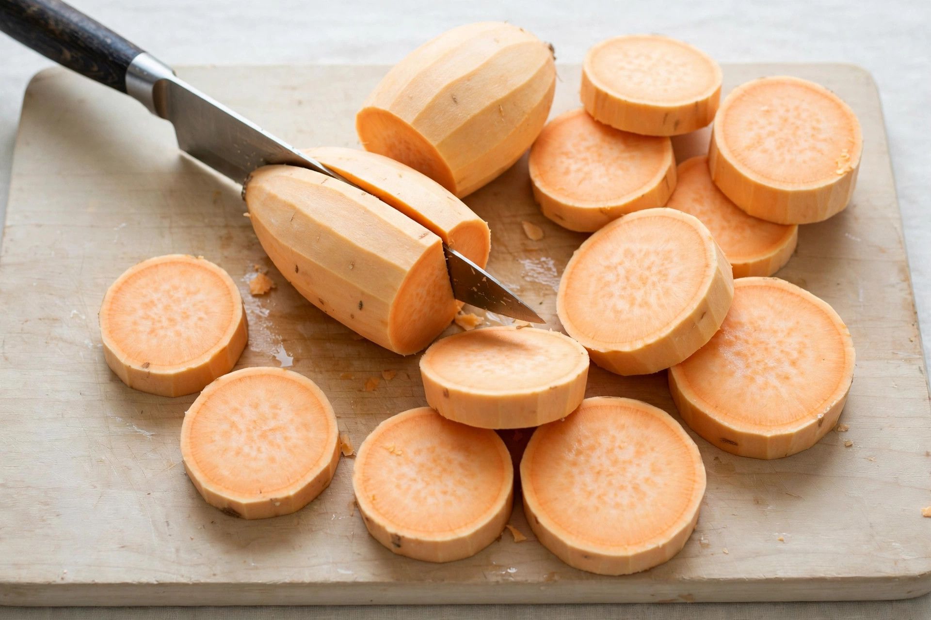 Slicing peeled sweet potatoes into thick even rounds on a wooden cutting board