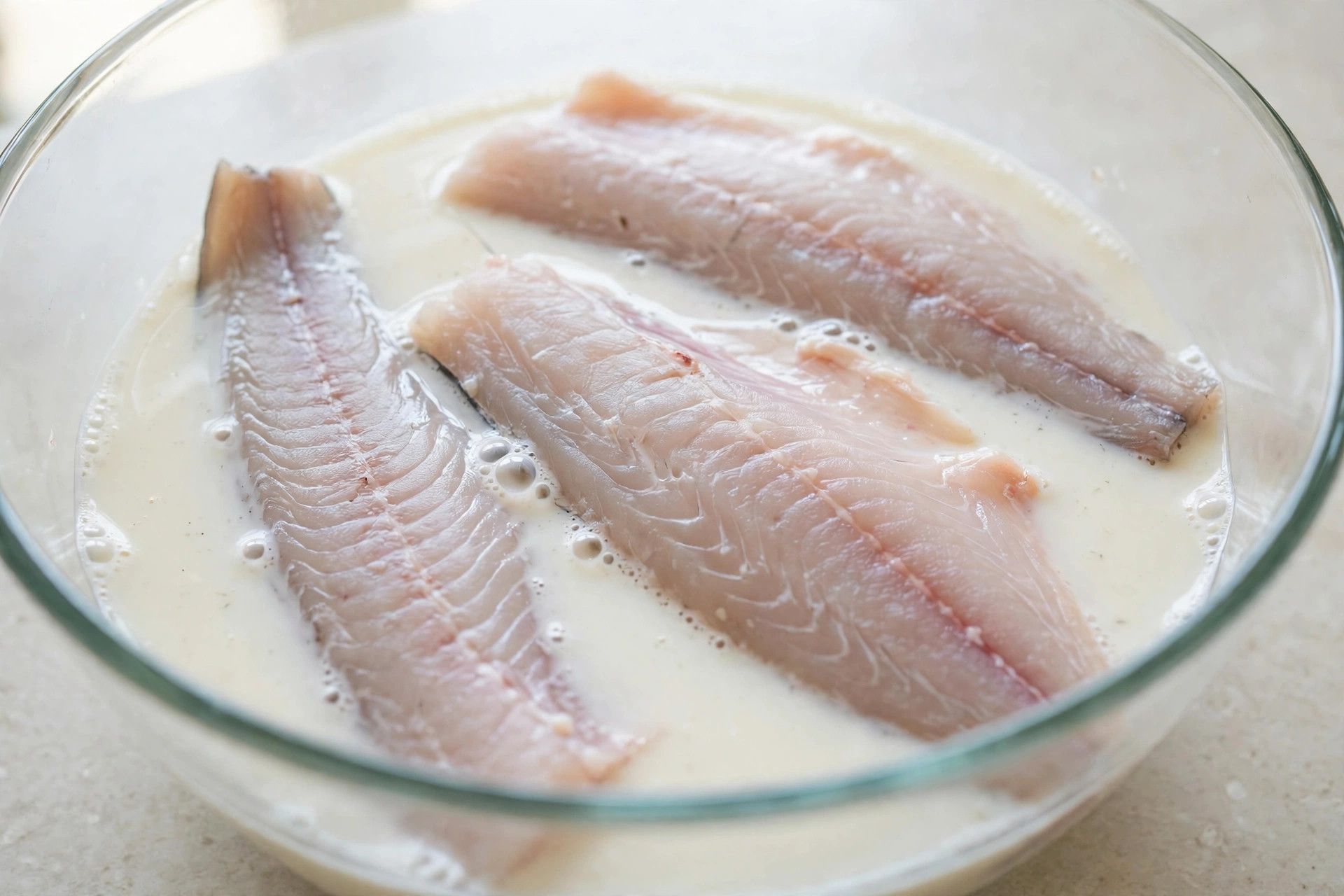 Raw catfish fillets soaking in buttermilk in a clear glass bowl on a light counter